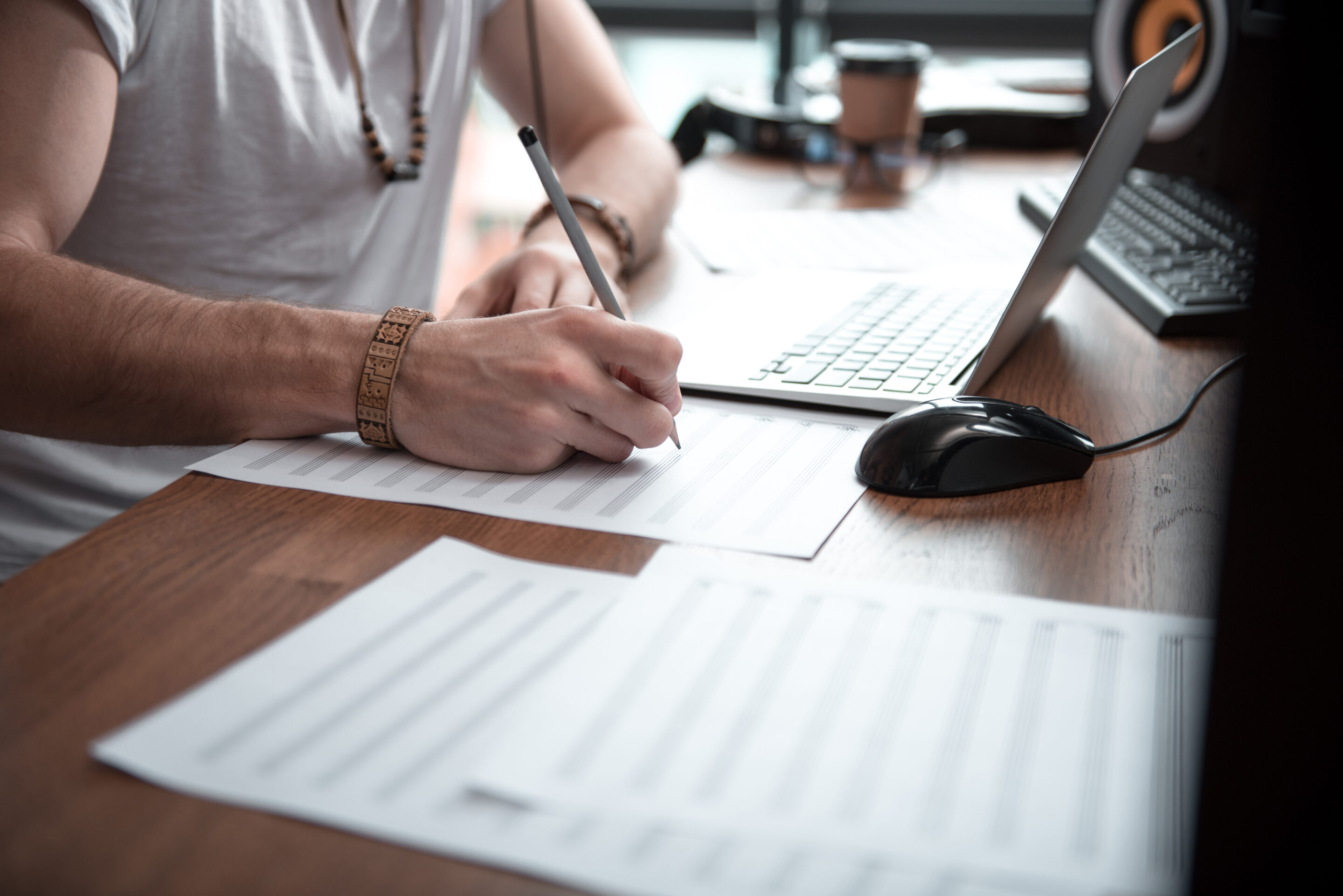 Close up of male hands drawing musical notes on paper. Man is sitting at desk near laptop.
