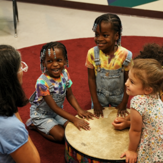 Three young children and a teacher around a large drum.