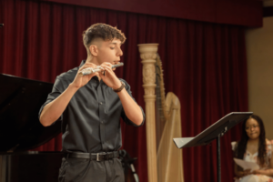 Teen boy playing the flute with a harp in the background.