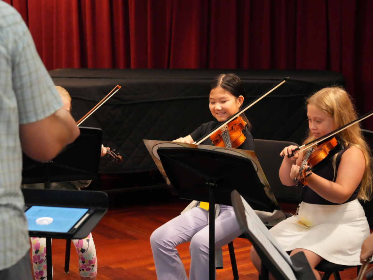 Two girls playing violin with a conductor in the foreground.